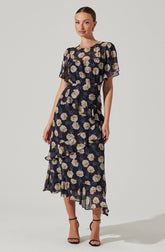 The woman is wearing a black dress with a floral pattern featuring large white, yellow, and purple flowers. The dress has short, flutter sleeves, a gathered waistline, and a tiered, ruffled hem that falls below the ankles. She is also wearing black high-heeled sandals.